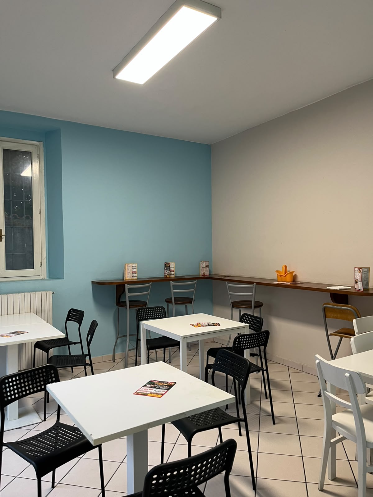 La saletta della pizzeria: tavolini bianchi, sedie nere e un bancone alto lungo la parete azzurra